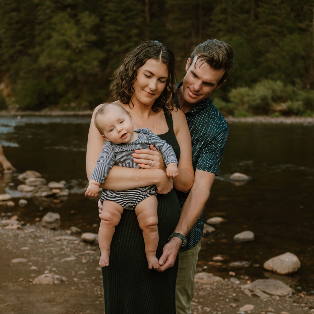 The Hickey family holding their baby, Charlotte, by a river surrounded by trees.
