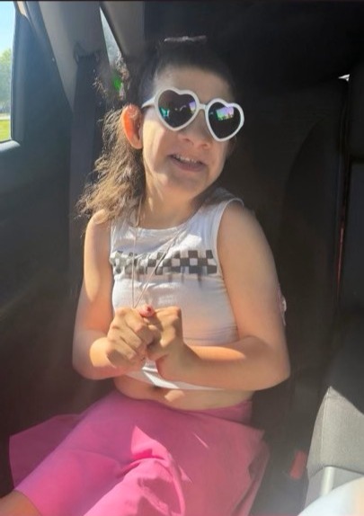 Nasyah Schwartz smiling in a car seat, wearing white heart-shaped sunglasses and a white top with pink pants.